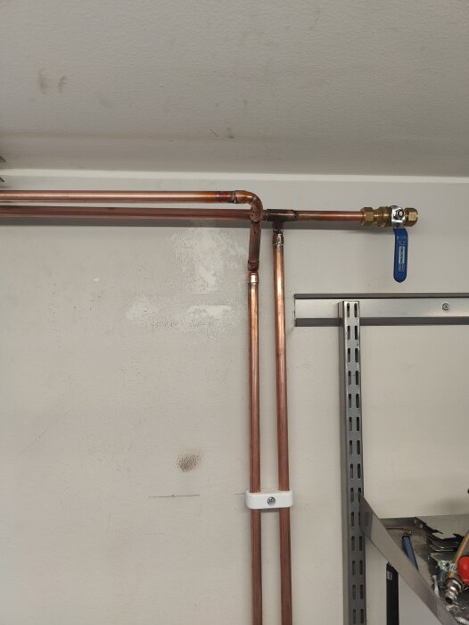 Kopparrörsledningar i garage med en 90-graders böj och blå ventil, installerade längs en vitmålad vägg, med hyllor och verktyg i bakgrunden.