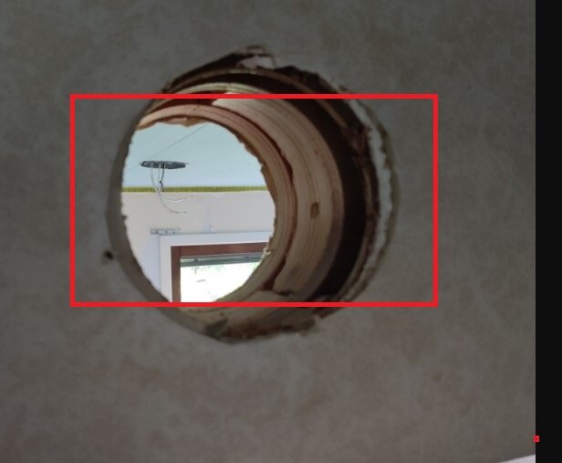 Rund håltagning i vägg visar träkonstruktion och en glimt av ett fönster och taklampa i bakgrunden. Potentiell plats för reparation med gipsskiva.