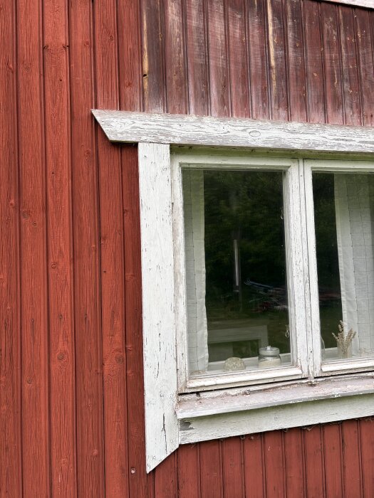 Röd träfasad med vita fönsterkarmar. Färgen är flagnad, och träet ser slitet ut. Ett fönster visar inredning med burkar och gardiner.
