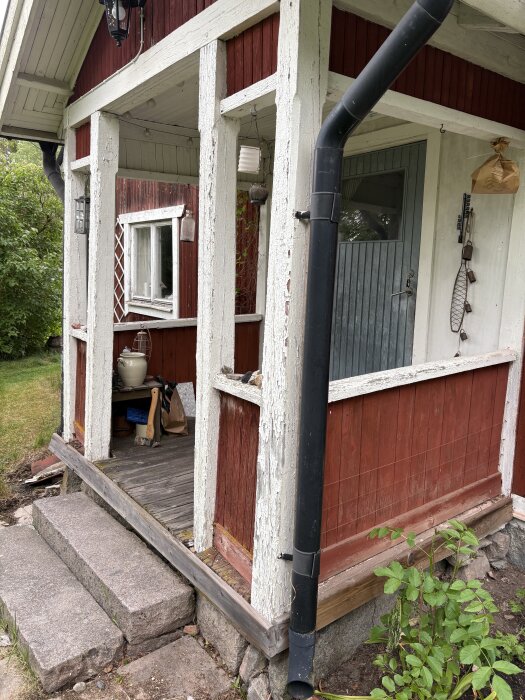 Gammal sommarstuga med rödmålad träfasad. Fönster, dörr och stuprör synliga. Träet är sprucket och delvis slitet. Stensteg leder till verandan.