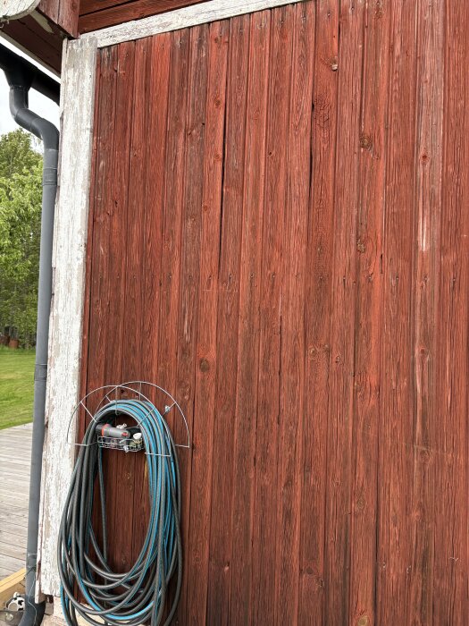 Gammal röd träfasad med sprickor, vitmålade hörn och en upphängd slang intill.