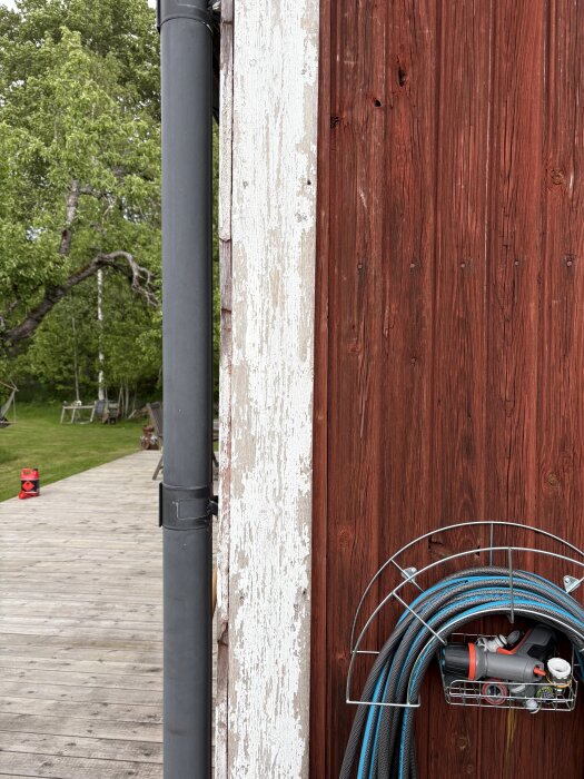 Träfasad på sommarhus med spruckna och avskalade delar, målad i falurött. Gårdsplan med träd och trätrall i bakgrunden. Hängd trädgårdslang.