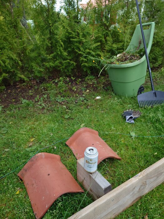 Öltunna med "Bitburger" på gräsmatta, omgiven av två tegelpannor och träd, med en grön vagn fylld med trädgårdsavfall i bakgrunden.