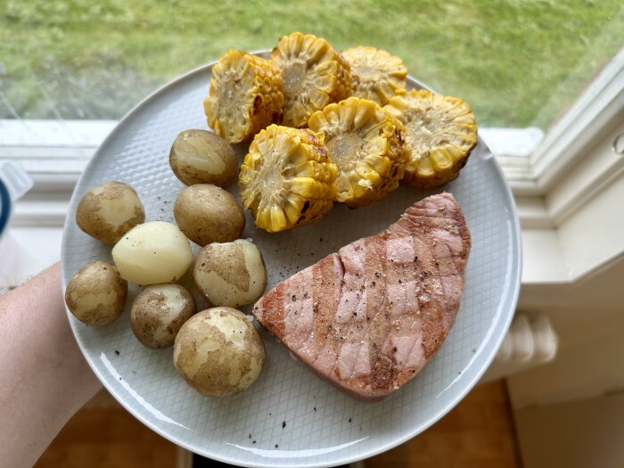 Tallrik med grillad tonfisk, färskpotatis och majskolvar på fönsterbräda, sommarkänsla.