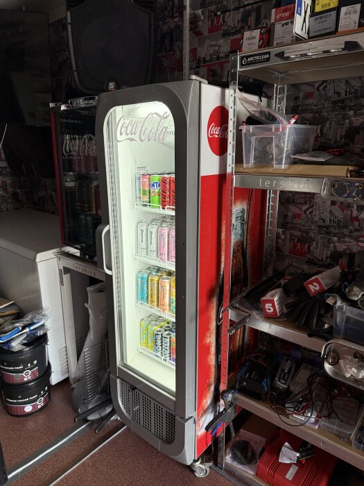 Coca-Cola kylskåp i garage med drycker; omgiven av verktyg och tillbehör på hyllor.