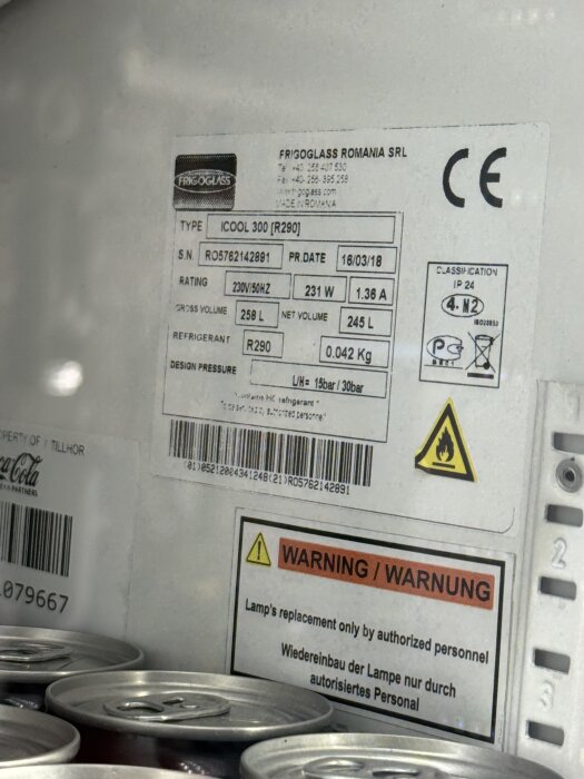 Etikett på ett Coca-Cola kylskåp med specifikationer och varningstext, stående på burkar, modell iCool 300 markeras.