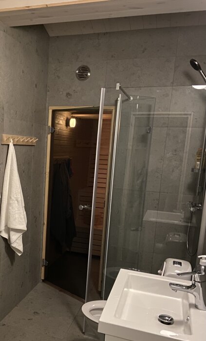 Badrum med klinkersvägg, glasad dusch och ingång till bastu. Handfat med kran syns till höger, handduk hänger på väggen.