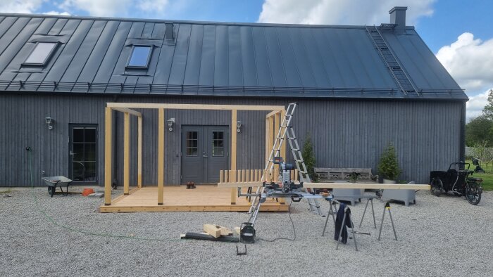 Pågående altanbygge med trästomme framför ett grått hus, verktyg och stege synliga, samt färdiga altandelar med nedre och övre bärlinor.