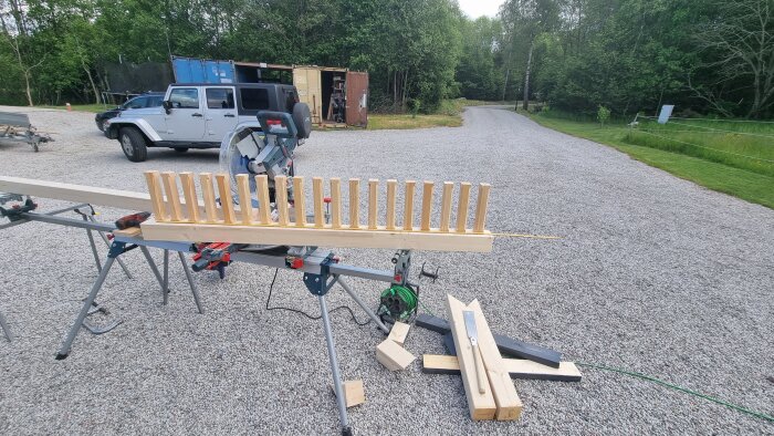 Trästolpar för trädäck, monterade på arbetsbänk utomhus. Verktyg och material syns runt omkring. Grönskande bakgrund.