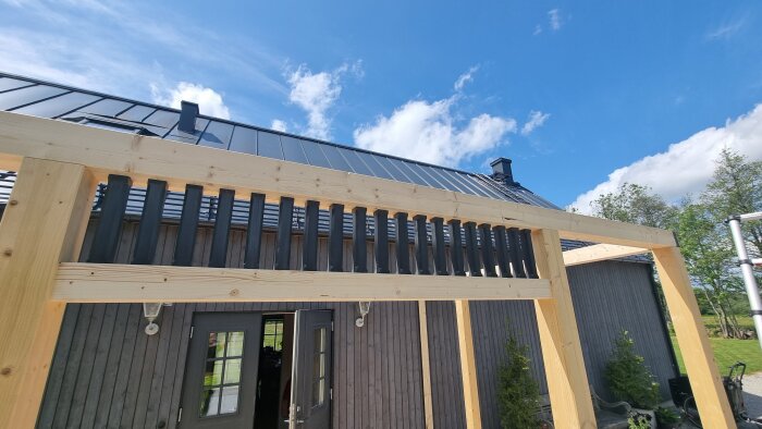 Träkonstruktion för trädäck under uppbyggnad framför grå fasad, med monterade stolpar och balkar. Blå himmel och grönska i bakgrunden.