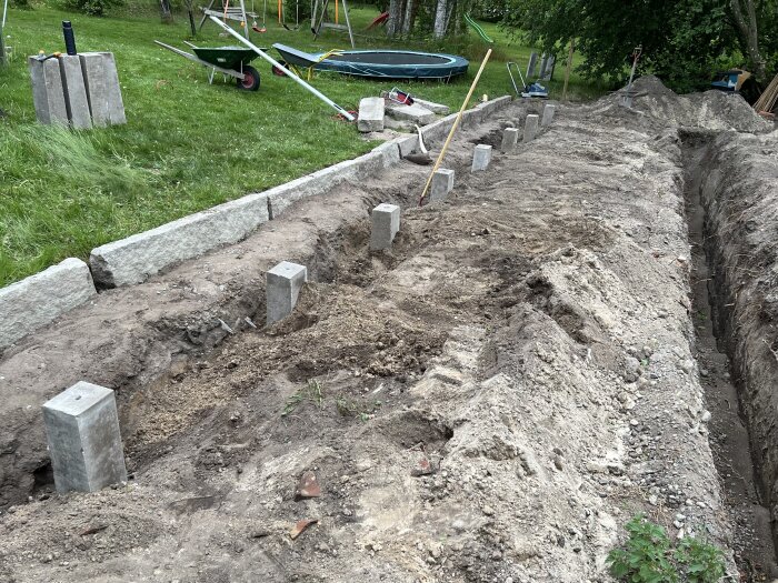 Rad av plintar på plats i en trädgård, med ölkar och grävmaterial bredvid, omgiven av gräsmatta och träd.