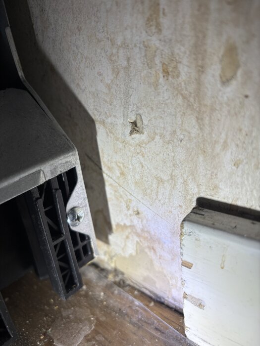 Vägg med synliga fläckar och skador bredvid hörnfäste och golvlist. Skuggor framhäver strukturen och eventuella målningsproblem.