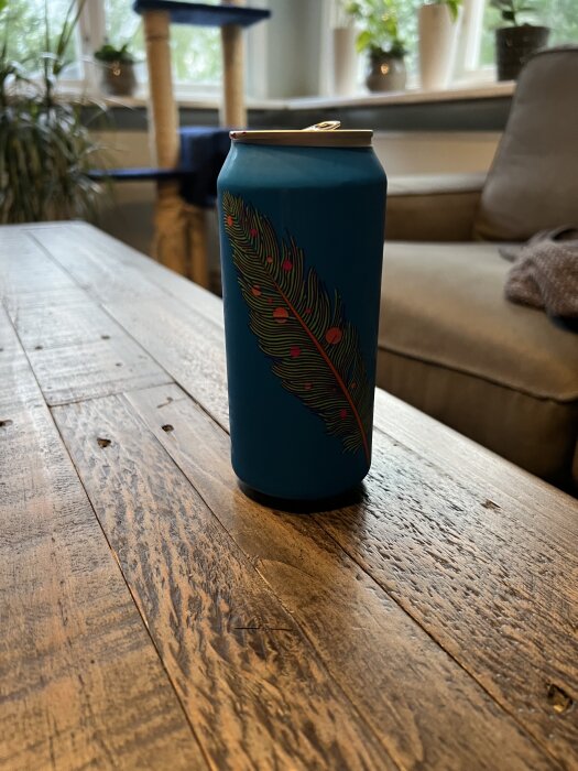 Blå dryckesburk med färgglatt fjädermotiv på rustikt träbord, omgiven av växter och inredning i bakgrunden.