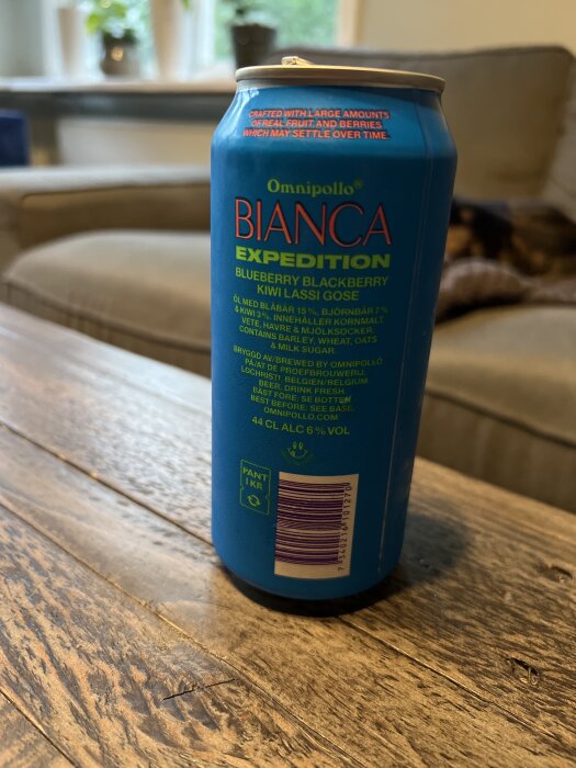 Blå burk med texten "Omnipollo Bianca Expedition", innehållande blåbär, björnbär och kiwi lassi gose, står på ett träbord i vardagsrumsmiljö.