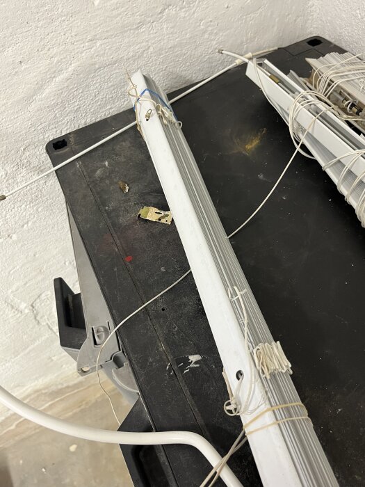 En persienn med en vit plåtbit och snören ligger på ett svart bord, med en vägg i bakgrunden.