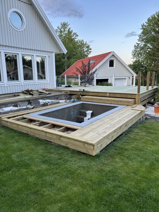 Träterrass med poolbygge på en gräsmatta, omringat av hus med vita fasader. Tralldäck delvis färdigställt.