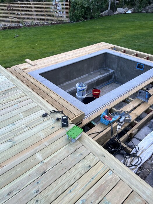 Trädäck med poolbygge; trallat övre däck, ofärdig pool med byggmaterial runtom, gräsmatta och staket i bakgrunden.