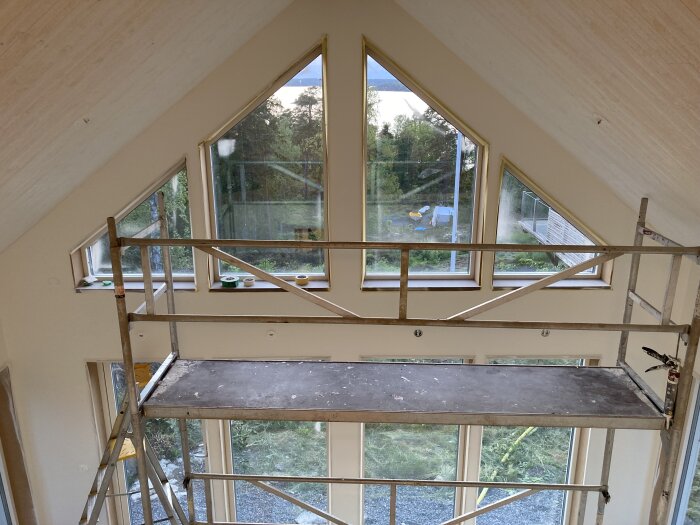 Ställningsplattform inomhus framför stora fönster i vinkel med utsikt över trädgård och parkering; renoveringsarbete pågår.
