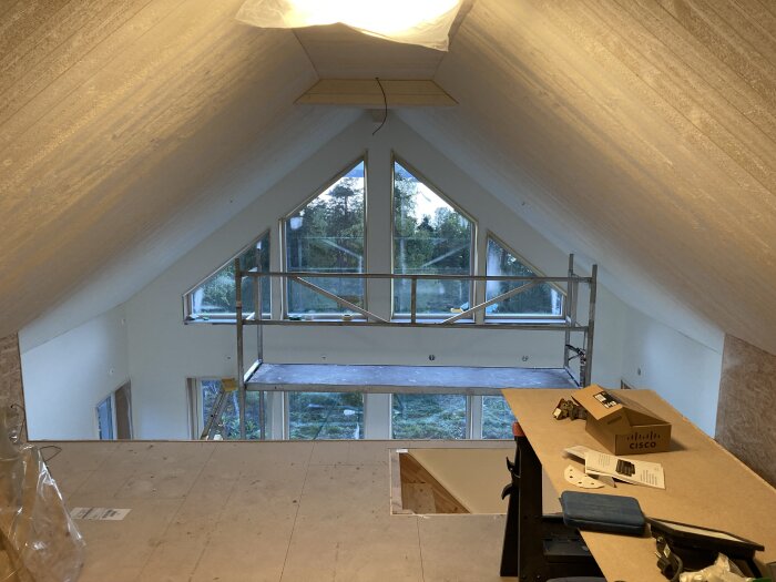 Ett rum under renovering med ett högt snedtak, stora trekantiga fönster, byggnadsställning och en Cisco-låda på ett bord.