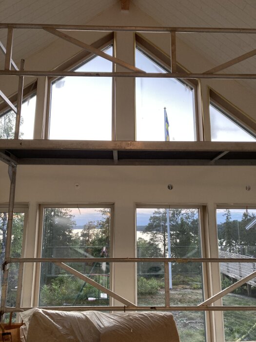 Interiör av hus under renovering med stora fönster och byggställning, utsikt över trädgård och svensk flagga utanför.