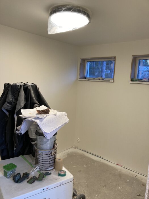Rum under renovering med målarburkar och verktyg på ett skåp, nyinstallerad taklampa och fönster som visar träd utanför.