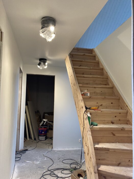 Trappor i trä och nymålade väggar i ett pågående renoveringsprojekt. Kablar och verktyg syns på trappan och golvet.