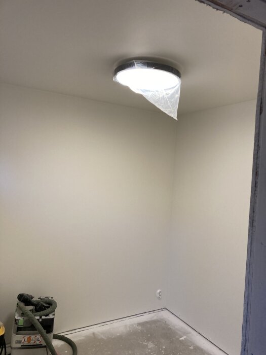 Vitmålat rum med taklampa täckt av plast. Verktyg och dammsugare syns i hörnet.