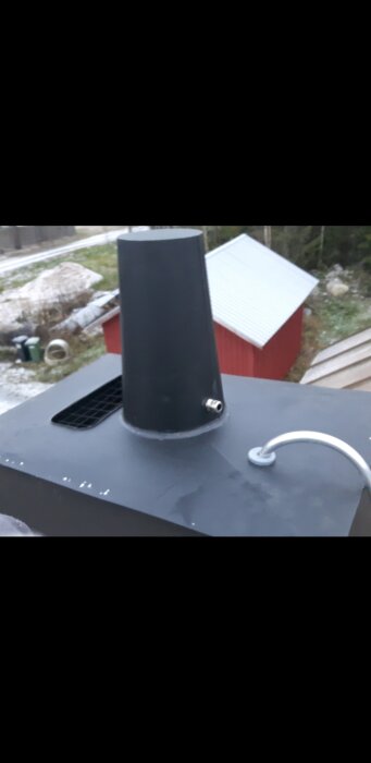 Svart Vilpe takfläkt monterad på ett tak, omgiven av små byggnader och träd i bakgrunden.