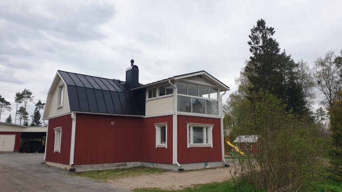 Rött hus med svart tak och Vilpe takfläkt installerad, omgiven av grönska och en mindre byggnad i bakgrunden.