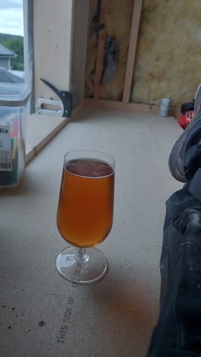 Ett glas med dryck på ett halvfärdigt träunderlag, en byggmiljö med delvis isolerad vägg i bakgrunden.
