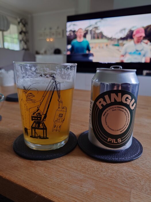 En pilsner i glas bredvid en burk med etiketten "Ringu Pils" på ett bord, TV i bakgrunden.