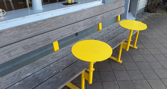 Träbänk med två runda gula bord på sidan av en trottoar framför ett fönster.