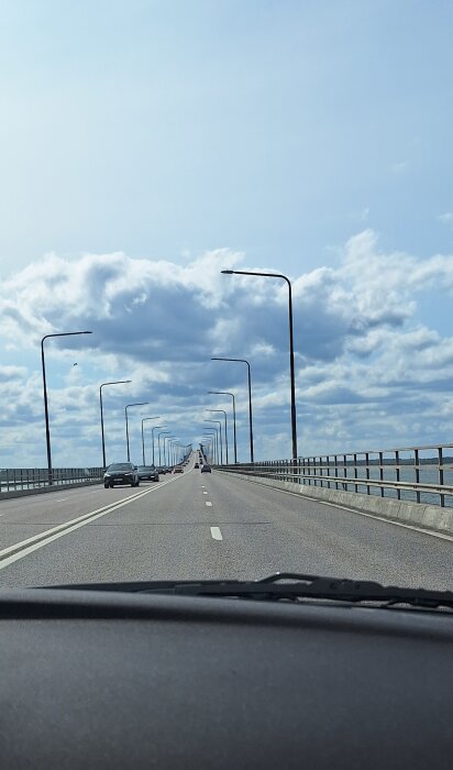 Vy från en bil över en lång bro med gatlampor, några bilar framför och molnig himmel ovan.