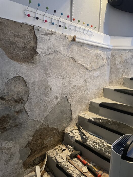Trasig källarvägg bredvid en trappa där gammalt spackel och puts avlägsnats, med verktyg och skräp på trappstegen.
