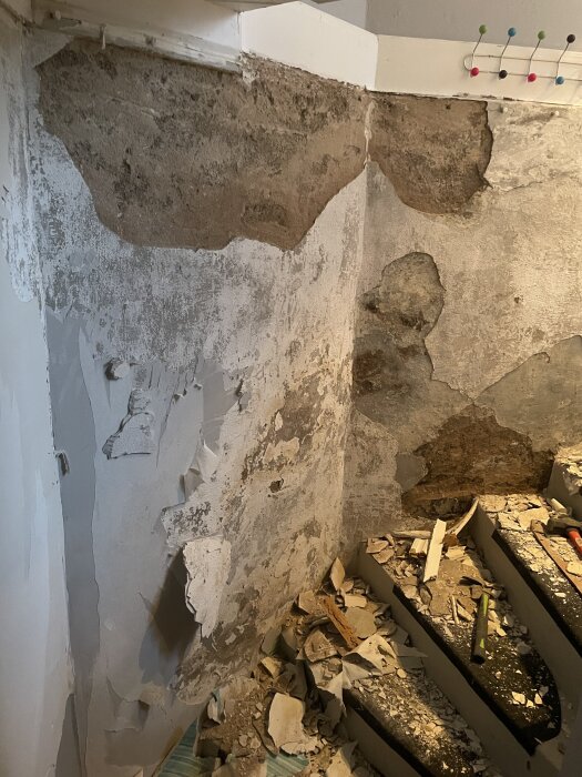 Uppbruten källarvägg med flagnande puts och synlig betong; byggavfall och verktyg ligger på en trappa bredvid väggen.