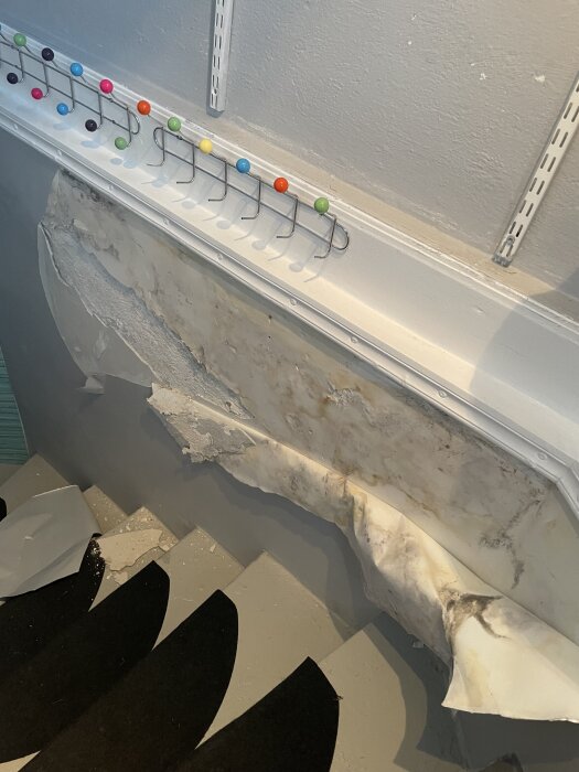 Skadad källarvägg med avflagad renoveringstapet och synlig betong bakom, omgiven av färgglada krokar på vägghylla.