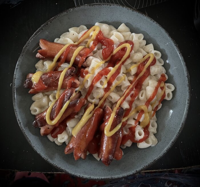 Stuvade makaroner med prinskorv, toppat med ketchup och senap på en tallrik.