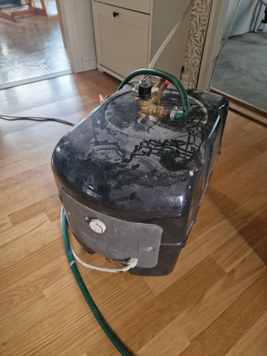 Tillfällig varmvattenberedare med gröna och silverfärgade slangar samt guldiga ventiler, placerad på trägolv i ett hem under renovering.
