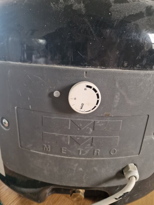 Närbild på en svart varmvattenberedare med en vit temperaturinställningsknapp, märkt "Metro". Synliga kablar och smuts på ytan.