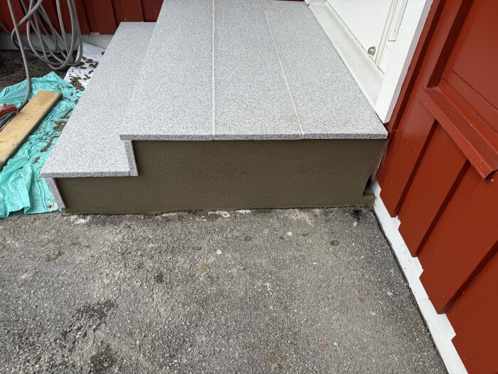 Steg vid entré med grå stenar och putsad cementbrukssida, röd vägg i bakgrunden.