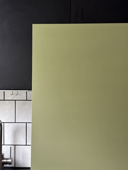 Grönt köksskåp under ommålning, nära motiv visar ojämn yta med "apelsinskal" textur, vitt kakel och svart bänkskiva i bakgrunden.