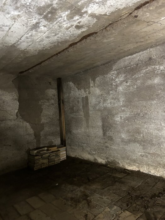 Fuktigt utrymme under balkong med rostigt armeringsjärn och fuktskadade reglar, staplade plattor och tydliga vattenfläckar på väggar och golv.