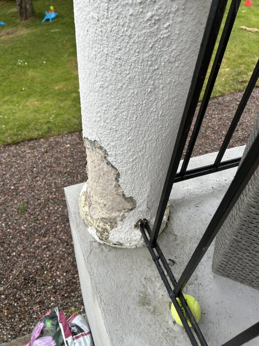 Skadad puts på en pelare utomhus med avskalat underlag, bredvid ett järnräcke och en tennisboll. Gräs och grus syns i bakgrunden.