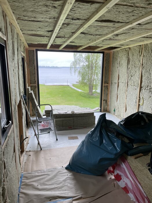 Byggarbetsplats, interiör med isolerade väggar och tak, stege och sopkasse. Vy mot sjö genom öppen vägg.