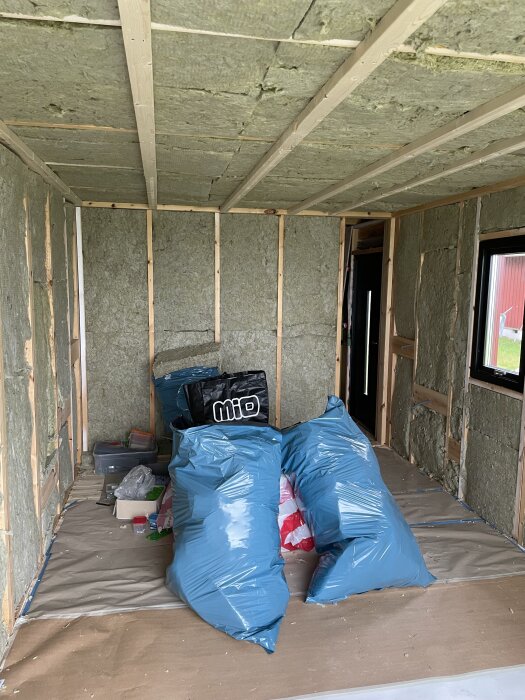 Isolerade väggar och golv i ett rum under renovering med byggmaterial i blå säckar på golvet.