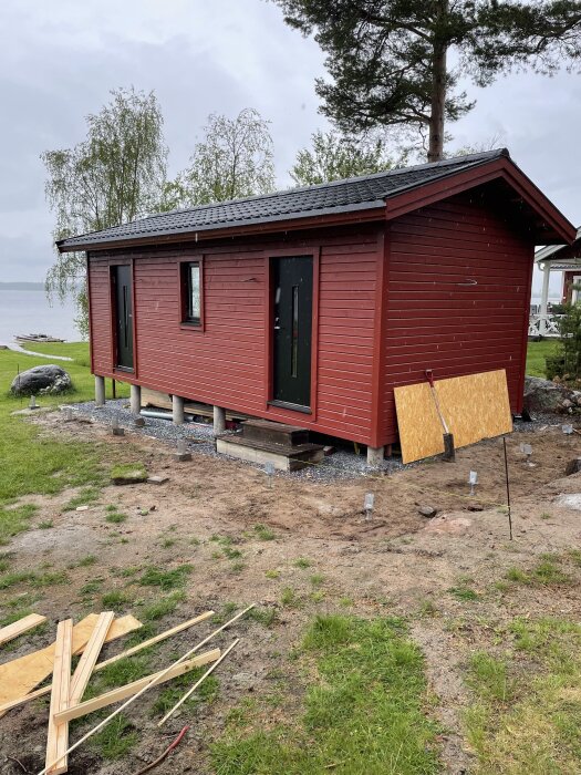 Byggnaden i trä vid sjön med nedgrävda plintar för altan, samt byggmaterial på marken.