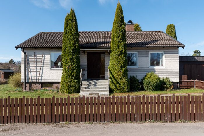 Hus med källare, omgiven av buskar och träd med en dränering från 90-talet, inramat av staket och trädgård i förgrunden.