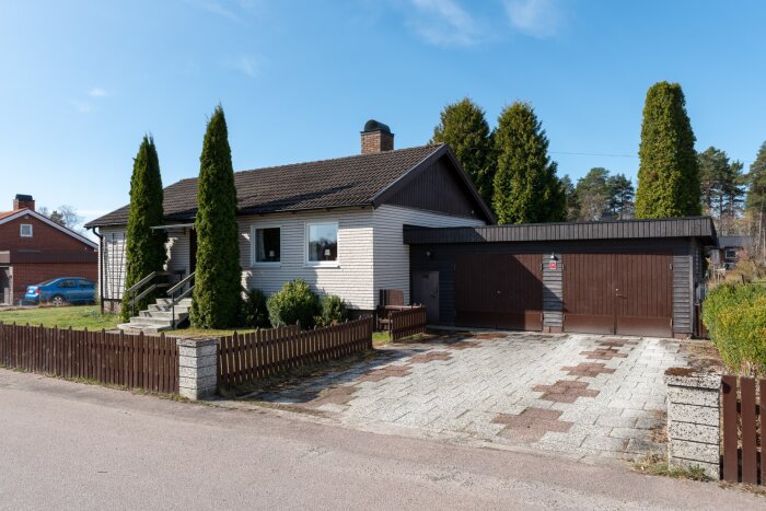 Ett hus med flera buskar och träd nära fasaden och en garageuppfart, omgett av ett litet staket och en bit bort ses en bil.