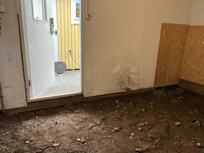 Dörröppning i garage med jordig golvyta och synlig byggdörr. Utsidan visar träterrass och gul vägg.