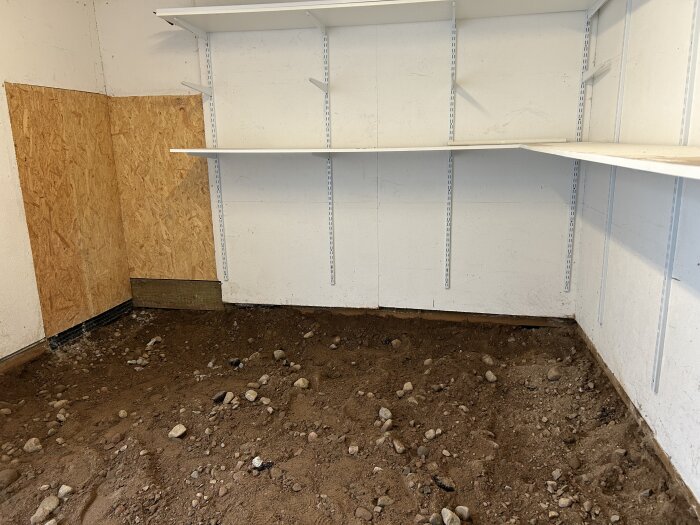 Uppgrävt garagegolv med jord och stenar, vita hyllor på väggen och en träpanel i hörnet.